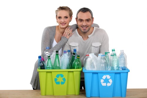 A young woman and a young man are positioned behind two brightly coloured recycling bins placed on a wooden table, with a plain white background. The woman has blonde hair styled in an updo, is wearing a light grey cardigan over a white top, and has her hand gently resting on the man's shoulder. The man has short dark hair, is dressed in a light grey long-sleeved shirt, and is smiling at the camera. The green recycling bin in front of them contains clear plastic bottles, some with labels, and a few crushed, arranged loosely. The blue recycling bin next to it contains similar clear plastic bottles and containers, also loosely arranged. Both bins are made of plastic, with a characteristic recycling symbol printed on their fronts in white, indicating their purpose for waste segregation. The scene conveys a clean, environmentally-conscious approach to waste management within a domestic or small-scale commercial setting, possibly reflecting services provided by Commercial Waste Removal Stoke Newington, operating in or near Stoke Newington, London. The overall atmosphere emphasizes recycling and waste separation, relevant to rubbish removal services.