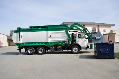 A large commercial rubbish collection truck, painted in white with green accents, is parked on a paved driveway in front of a modern residential building. The truck features a hydraulic arm mechanism extending from the front of the vehicle towards a blue skip located on its right side, which is positioned on the driveway. The skip appears to be filled with waste materials and has a weathered surface. The background includes a contemporary two-story house with white walls and a flat roof, situated in a quiet suburban area. The environment is well-lit, suggesting daytime with clear weather conditions. The truck is part of a waste management service, likely operated by Commercial Waste Removal Stoke Newington, servicing local rubbish collection needs within the Stoke Newington postcode area. The scene illustrates the process of waste collection, emphasizing the company's role in managing household and commercial refuse in the local community.