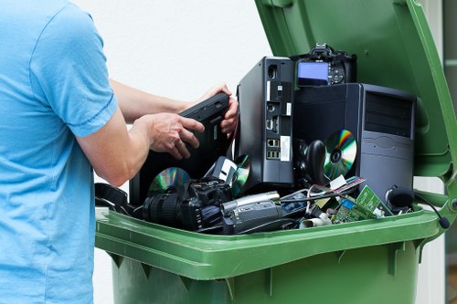 A person wearing a light blue shirt is seen using a handheld device over a green wheeled rubbish bin filled with electronic waste, including a computer tower, a monitor, CDs, and assorted cables and gadgets. The electronic waste occupies a significant portion of the open bin, which is located outdoors on a paved surface, with a background that suggests an urban or residential setting. The devices exhibit various textures, with the computer tower having a matte black finish and the CDs reflecting light. The scene is well-lit with natural daylight, emphasizing the details of the electronic components. This image relates to waste management services, such as those provided by Commercial Waste Removal Stoke Newington, that handle the collection and disposal of electronic and general rubbish in the Stoke Newington area, highlighting proper electronic waste recycling or removal procedures.