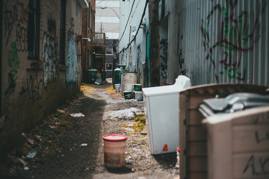 A small, silver aluminum step ladder with metal rails and a textured platform in the middle stands upright on a gray concrete pavement within a narrow outdoor space. The ladder is positioned against a