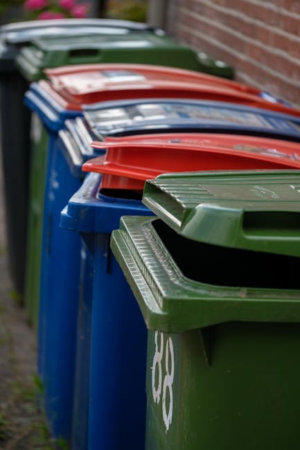 A row of large, outdoor wheelie bins lined up along a pavement adjacent to a brick wall. The bins are primarily blue with a green bin at the front and red, green, and blue lids on top. The lids are fl